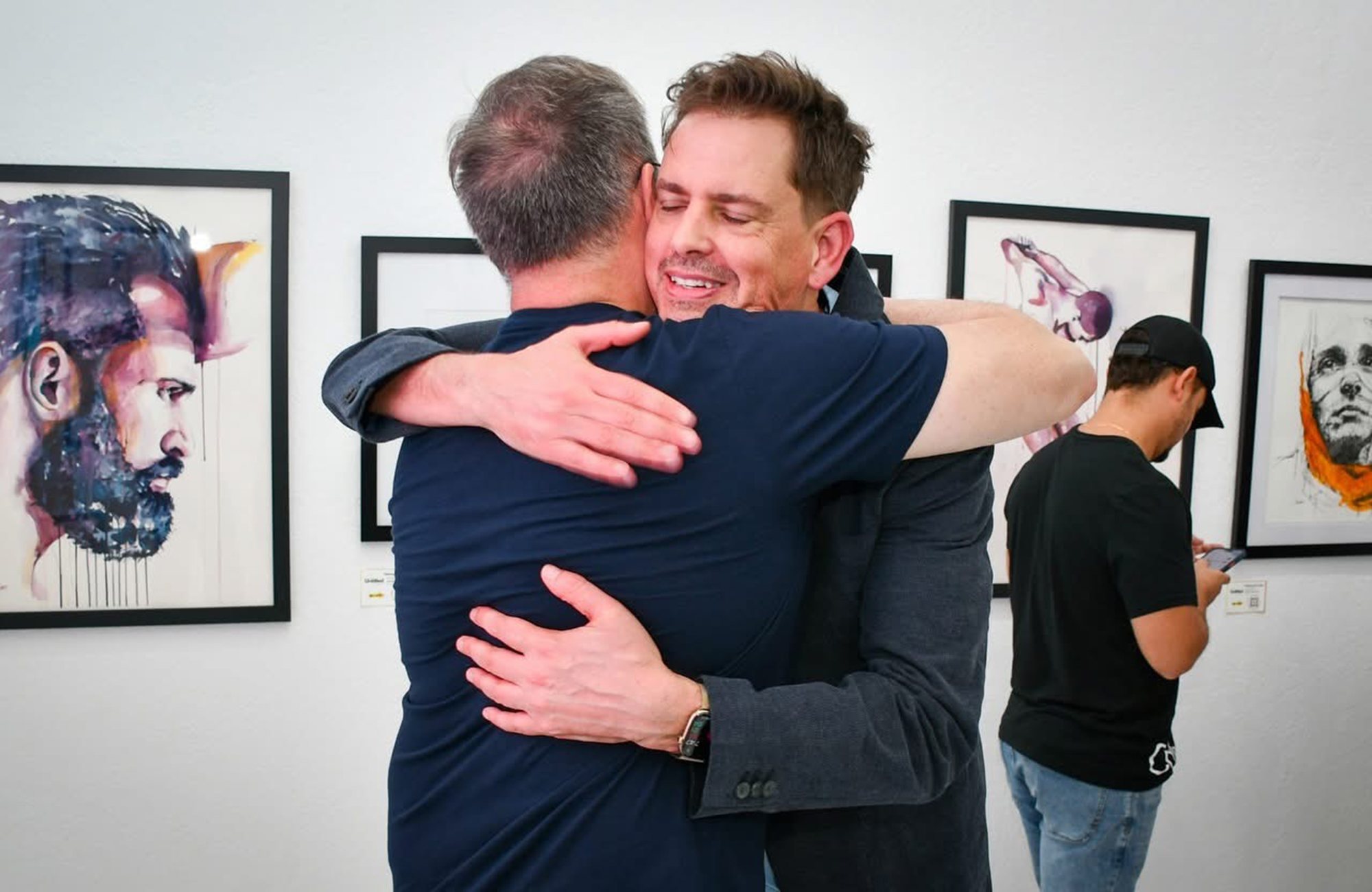 Brenden Sanborn sharing a hug at an art opening, with his watercolor portraits of men hanging on the gallery wall behind them