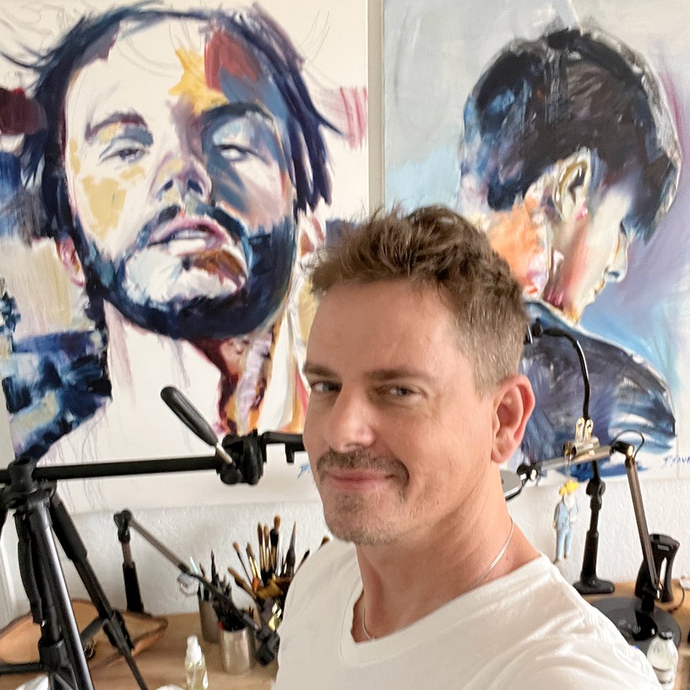 Artist Brenden Sanborn smiling in his studio in front of two large expressive portrait paintings