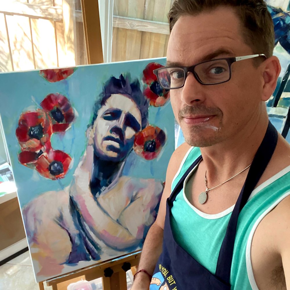 Brenden Sanborn in his apron taking a selfie in the studio beside his painting of a man with red poppies on the easel
