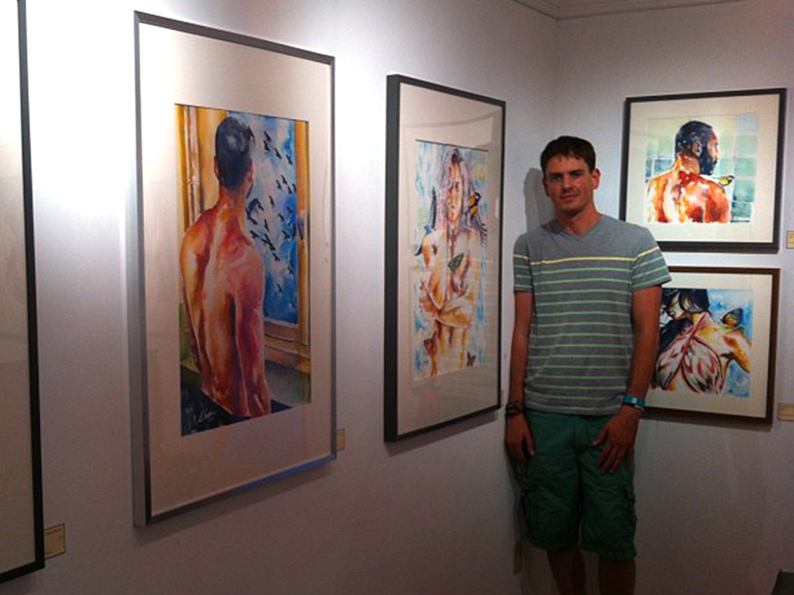 Brenden Sanborn standing beside his framed watercolor paintings of male figures at a gallery show