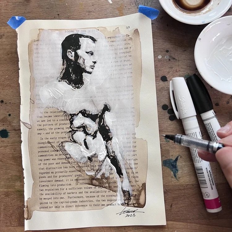 Brenden Sanborn's custom ink portrait on a book page with paint markers and coffee on his studio table
