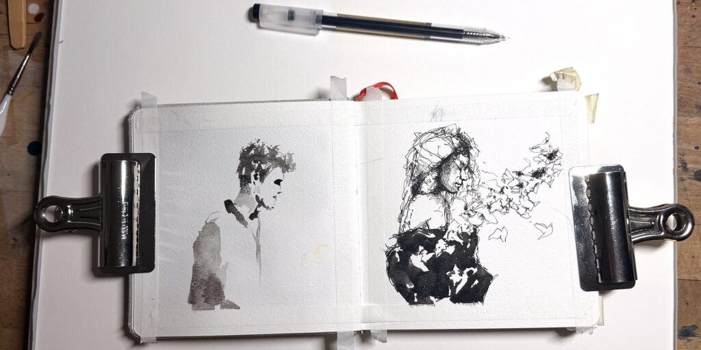 Open sketchbook in Brenden Sanborn’s studio with two expressive ink figure drawings held flat by binder clips
