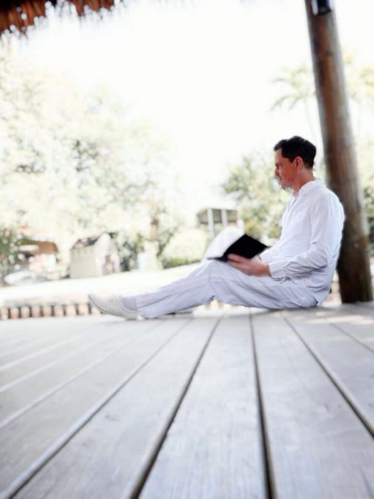 Brenden Sanborn in white sitting on a wooden deck outdoors with an open sketchbook