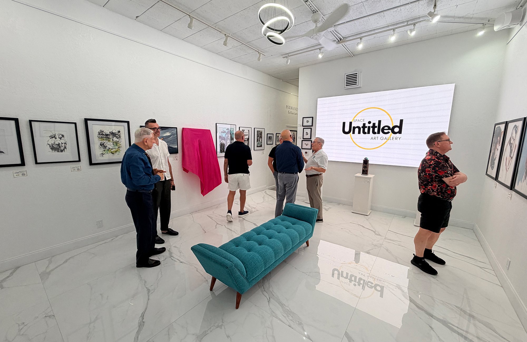 Guests at an art opening at Space Untitled Art Gallery in Wilton Manors, Florida, with framed works on the walls and the gallery logo on a large screen