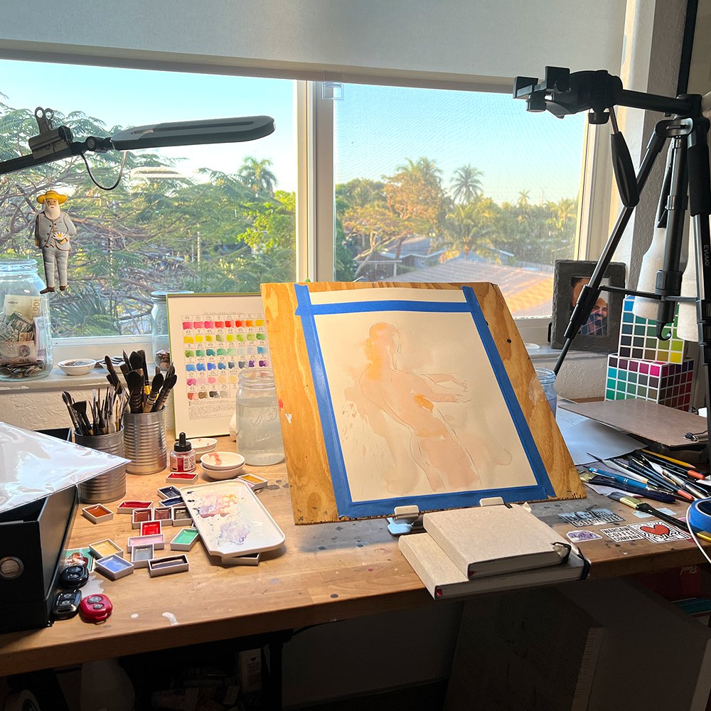 Brenden Sanborn watercolor studio desk by a sunny window with a taped watercolor on a board, brushes and paints