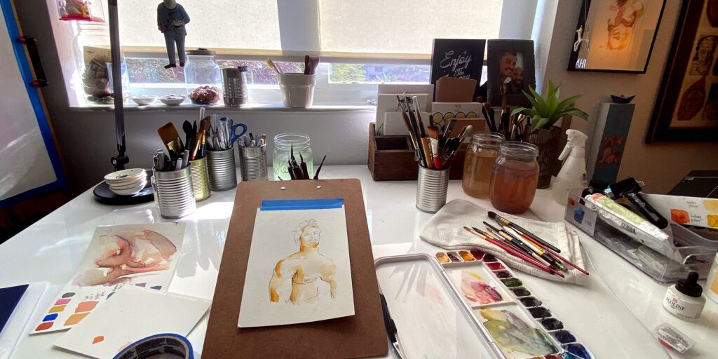 Brenden Sanborn’s studio desk with a male figure watercolor in progress on a clipboard, brushes, paint jars and a watercolor palette by the window