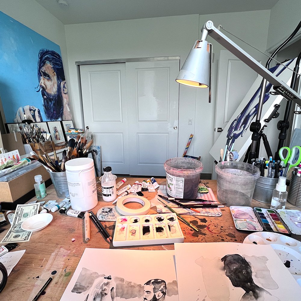 Brenden Sanborn studio desk covered in brushes, ink and watercolor paints, with a large blue portrait painting on the wall