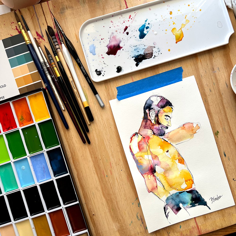 Watercolor palette, brushes and a watercolor figure painting of a bearded man on a wooden studio table