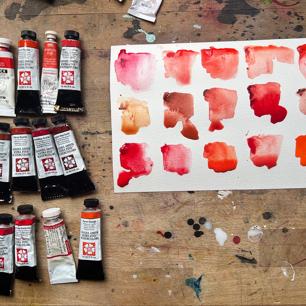 Red and orange watercolor color swatches beside tubes of Daniel Smith watercolor paint on a paint splattered table