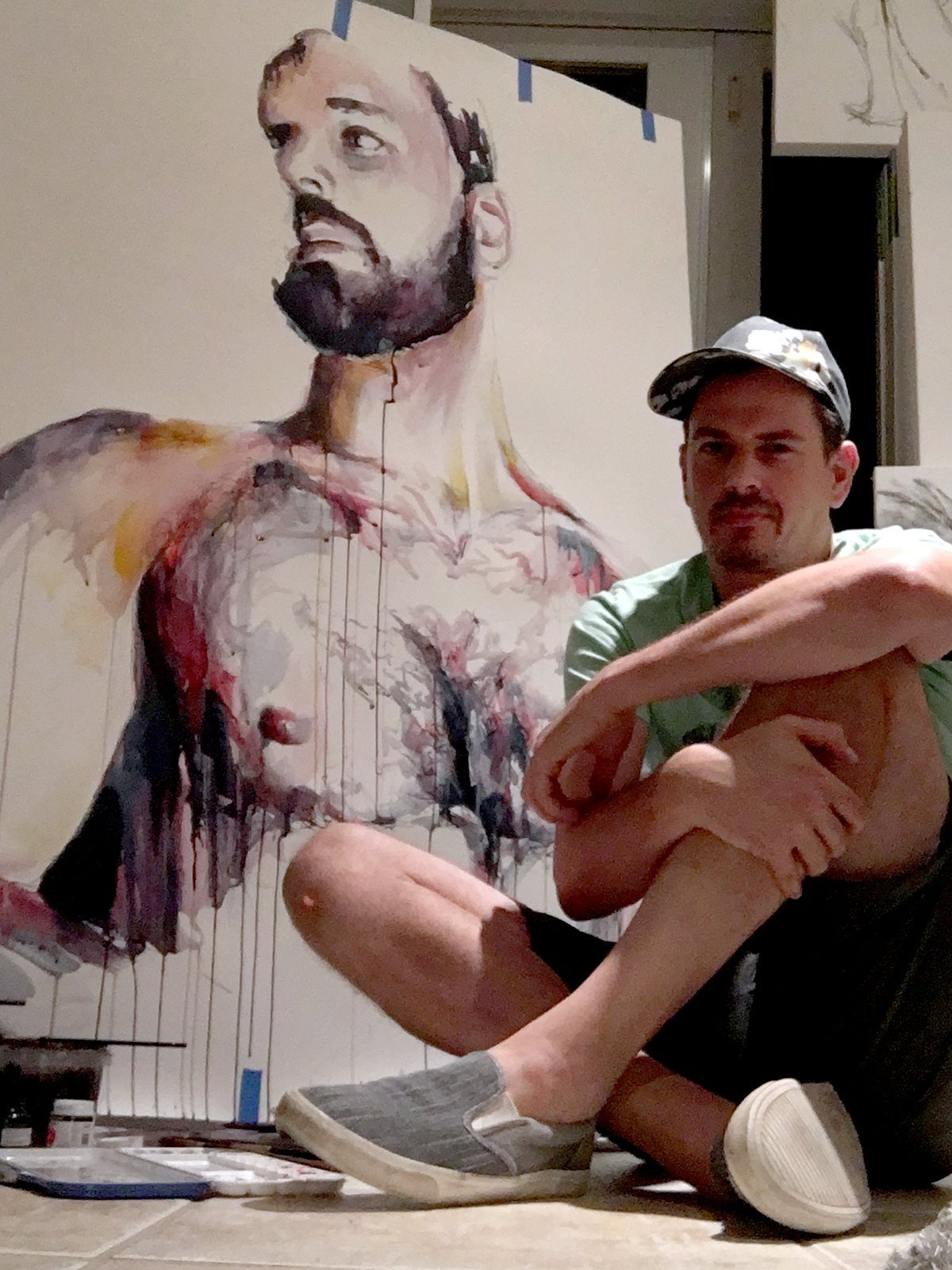 Brenden Sanborn sitting on the studio floor in front of a large watercolor portrait of a bearded man